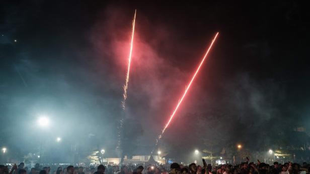 Viele Menschen stehen nachts im Freien und beobachten zwei aufsteigende rote Feuerwerksraketen.