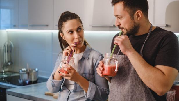 Zwei Personen trinken gemeinsam einen roten Smoothie aus Gläsern mit Strohhalmen in einer modernen Küche.