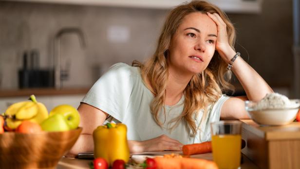Eine Frau sitzt nachdenklich am Küchentisch mit Obst, Gemüse, einem Glas Saft und einer Schale Mehl vor sich.