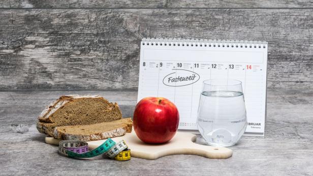 Ein Apfel, ein Glas Wasser, Brot, ein Maßband und ein Kalender mit dem Wort „Fastenzeit“ auf einem Holztisch.