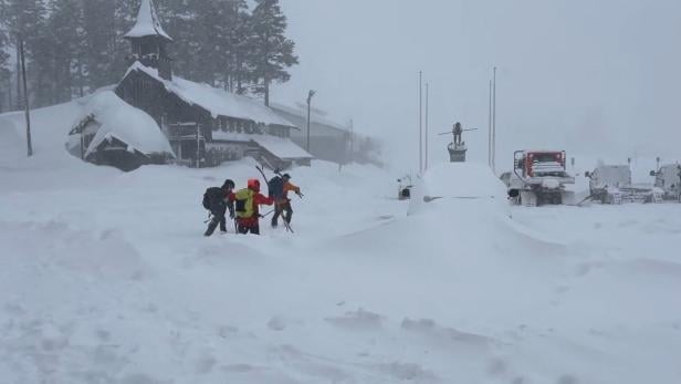 Mehrere Menschen mit Skiausrüstung kämpfen sich durch tiefen Schnee vor einem verschneiten Gebäude und Fahrzeugen.