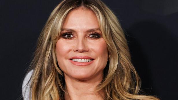 Heidi Klum mit langen blonden Haaren trägt ein tief ausgeschnittenes rotes Kleid und lächelt vor dunklem Hintergrund.
