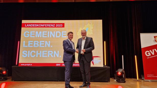 Zwei Männer im Anzug geben sich auf einer Bühne vor einem großen Bildschirm mit Konferenzschriftzug die Hand.