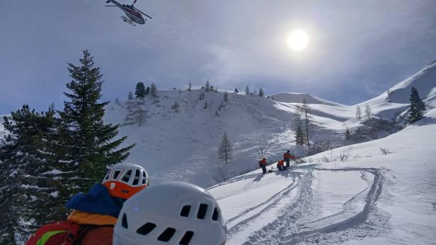 Ein Hubschrauber fliegt über eine verschneite Berglandschaft, während Bergretter zu sehen sind.