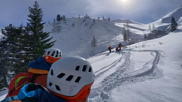 Ein Hubschrauber fliegt über eine verschneite Berglandschaft, während Bergretter zu sehen sind.