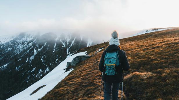 Eine Wanderin mit Winterkleidung und Rucksack geht auf einem verschneiten Berghang, umgeben von Nebel und karger Landschaft.