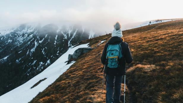 Eine Wanderin mit Winterkleidung und Rucksack geht auf einem verschneiten Berghang, umgeben von Nebel und karger Landschaft.
