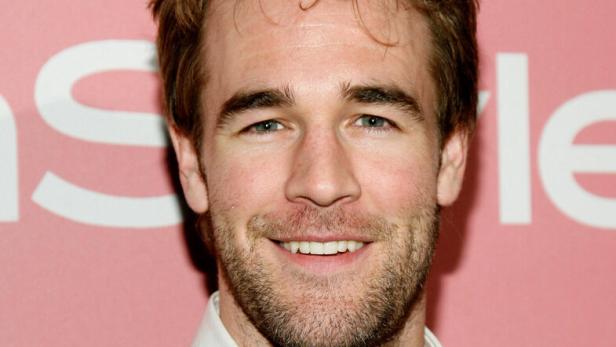 Ein lächelnder James Van Der Beek mit Bart und grauem Pullover vor rosafarbenem Hintergrund.