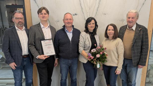 Sechs Personen stehen nebeneinander, eine Person hält eine Urkunde, eine andere einen Blumenstrauß.