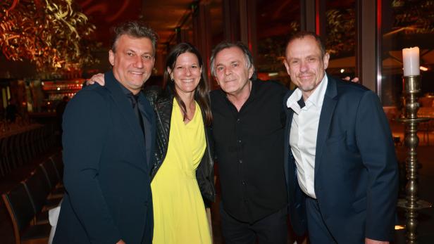 Vier Personen stehen eng beieinander und lächeln in eleganter Abendkleidung in einem festlich beleuchteten Raum.