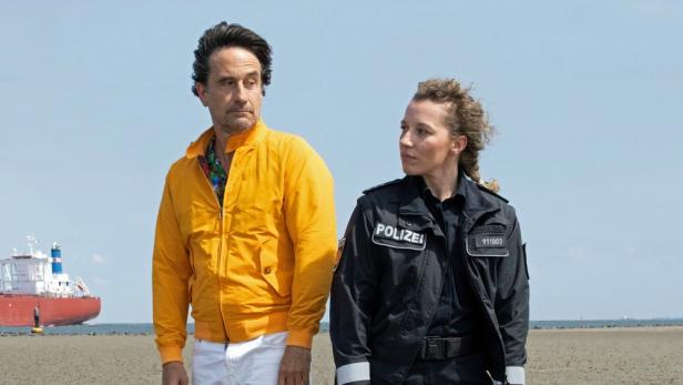 Frau in Polizeijacke und Mann in oranger Jacke blicken einander auf einem Strand an