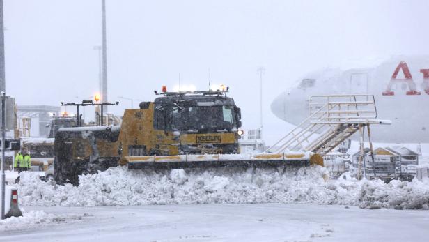 Ein gelber Schneepflug räumt Schnee auf einem Flughafen, im Hintergrund ein Flugzeug.