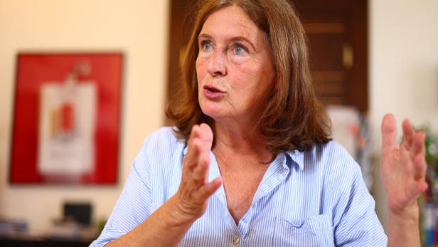 Eine Frau mit braunen Haaren und blau gestreifter Bluse gestikuliert beim Sprechen.