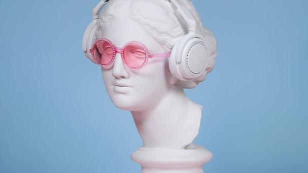 Eine weiße Büste trägt pinke Sonnenbrille und weiße Kopfhörer vor blauem Hintergrund auf rosa Fläche.