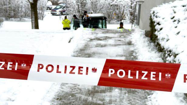 Ein rot-weißes Polizei-Absperrband versperrt einen verschneiten Weg, im Hintergrund sind Einsatzkräfte und ein Polizeiauto zu sehen.