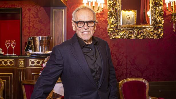 Wolfgang Puck mit Brille und dunklem Anzug steht in einem opulenten, rot dekorierten Raum mit goldenem Spiegel.