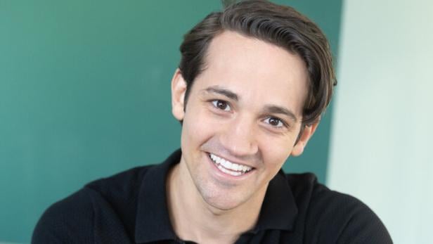 Ein junger Mann mit dunklem Haar und schwarzem Poloshirt sitzt lächelnd auf einem Stuhl und trägt eine weiße Armbanduhr.