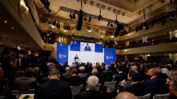 Ein Redner spricht vor einem großen, aufmerksam lauschenden Publikum in einem modernen Konferenzsaal mit mehreren Ebenen.