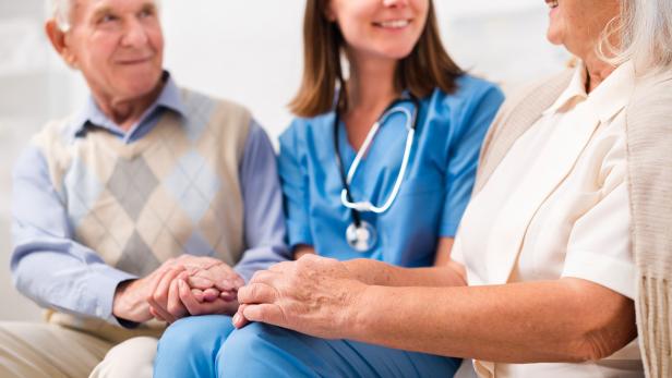 Pflegekraft im Gespräch mit älterem Paar, alle lächeln und halten sich an den Händen.