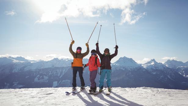 Drei Skifahrer stehen auf einer verschneiten Bergspitze und heben ihre Skistöcke in die Luft.