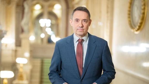 Ein Mann im Anzug mit roter Krawatte steht in einem prunkvollen, hell erleuchteten Innenraum.