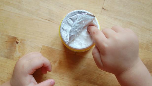 Ein Kind taucht einen Finger in eine geöffnete Dose mit weißer Creme auf einem Holztisch.