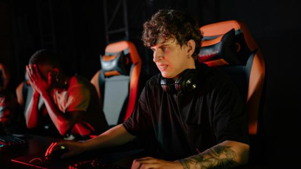 Zwei junge Männer sitzen an Gaming-PCs, einer wirkt frustriert, der andere konzentriert.