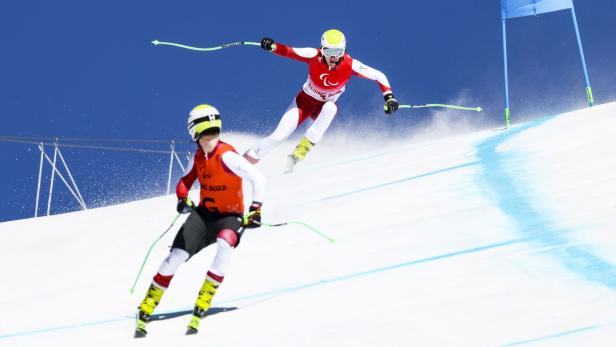 Zwei Skifahrer mit Helmen und Stöcken fahren eine steile, schneebedeckte Piste bei strahlend blauem Himmel hinab.