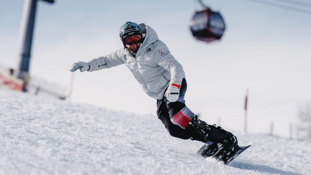 Der Para-Snowboarder Rene Eckhart mit Helm und Skibrille fährt auf einer schneebedeckten Piste, im Hintergrund eine Gondel.