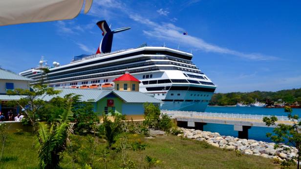 Ein großes Kreuzfahrtschiff liegt an einem sonnigen Hafen, während Passagiere durch eine bunte Hafenanlage mit kleinen Gebäuden an Land gehen.