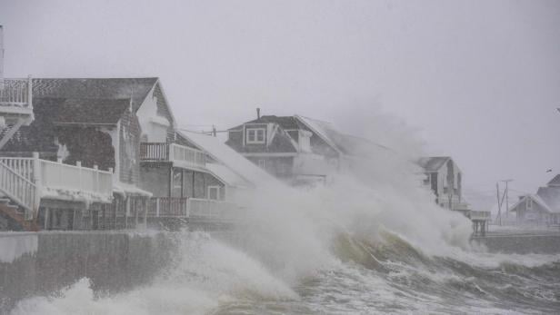Hohe Wellen schlagen gegen verschneite Häuser an einer Küste bei stürmischem Winterwetter.
