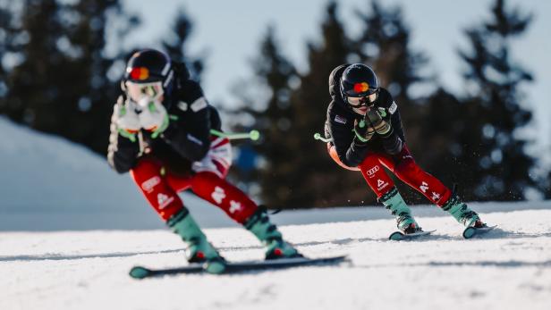 Die Skifahrerinnen Veronika und Elisabeth Aigner in Rennposition fahren schnell eine verschneite Piste hinunter, im Hintergrund unscharfe Bäume.