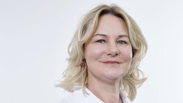 Frau mit blonden, schulterlangen Haaren trägt ein weißes Poloshirt mit Logos und lächelt leicht vor hellem Hintergrund.