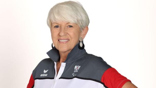 Maria Rauch-Kallath trägt Sportkleidung in Weiß, Grau und Rot und lächelt mit den Händen in der Hüfte.