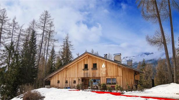 Holzhaus im verschneiten Wald mit Bergen im Hintergrund und einem roten Weg im Schnee vor dem Eingang.