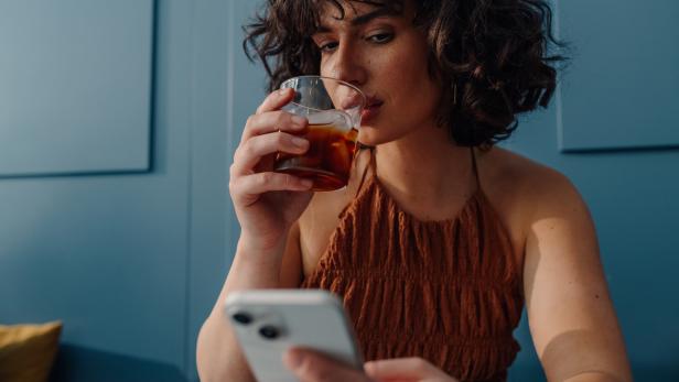 Eine Frau mit lockigem Haar trinkt etwas aus einem Glas und schaut auf ihr Handy.