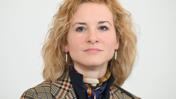 Frau mit blond gelockten Haaren, kariertem Blazer und Halstuch schaut ernst in die Kamera.