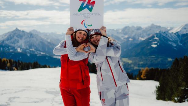 Veronika und Elisabeth Aigner stehen im Schnee vor Bergen und halten ein Schild mit dem Logo der Milano Cortina 2026 Paralympics.