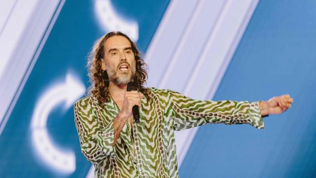 Russell Brand mit langen Haaren und gemustertem Hemd spricht in ein Mikrofon vor blauem Hintergrund.