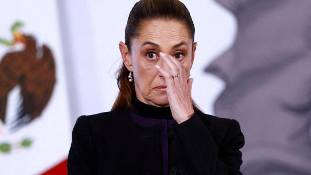 Eine Frau mit ernster Miene wischt sich mit der Hand über das Gesicht, im Hintergrund ist ein Teil einer Flagge zu sehen.