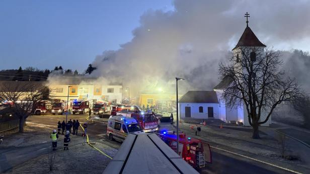 Dichter Rauch steigt aus einem brennenden Gebäude neben einer Kirche auf, zahlreiche Einsatzfahrzeuge und Feuerwehrleute sind vor Ort.