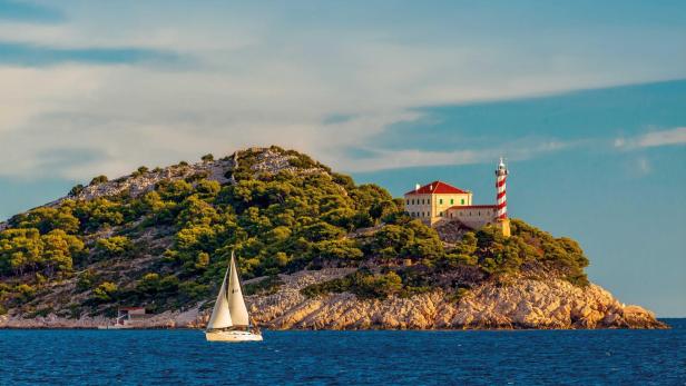 Eine kleine Insel mit grünem Bewuchs, einem rot-weißen Leuchtturm, einem Haus und einem Segelboot im Vordergrund.