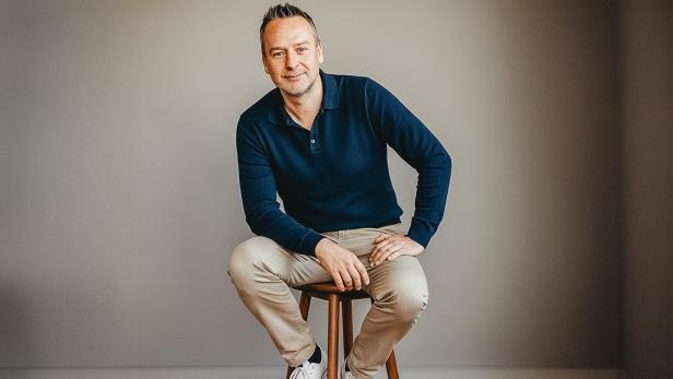 Mann in dunkelblauem Pullover sitzt entspannt auf einem Hocker vor neutralem Hintergrund.