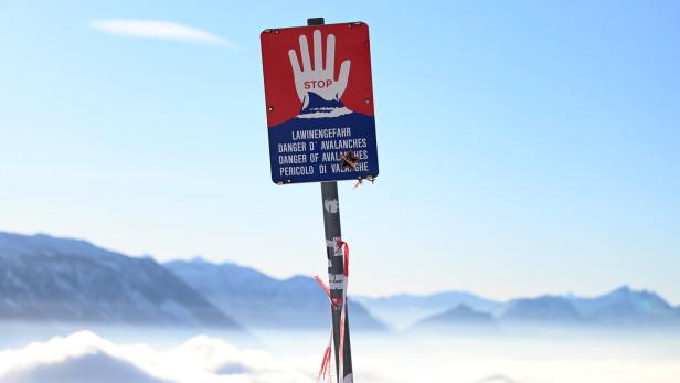 Warnschild mit roter Hand und Hinweis auf Lawinengefahr vor verschneiten Bergen und blauem Himmel.
