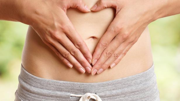 Zwei Hände formen ein Herz über dem Bauch einer Person, die eine graue Hose mit Kordelzug trägt.