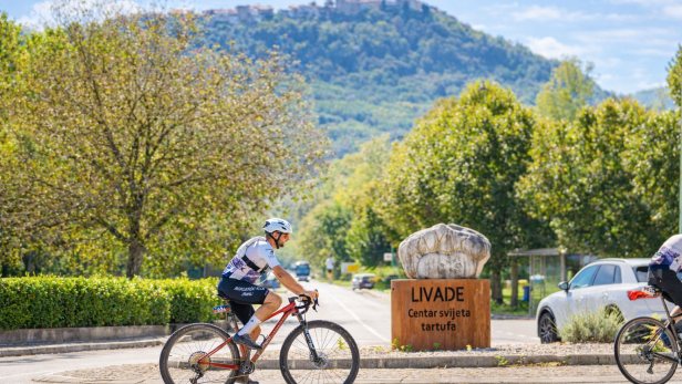 Radfahrer fahren an einem Schild für Livade vorbei, im Hintergrund ein Hügel mit Stadt.
