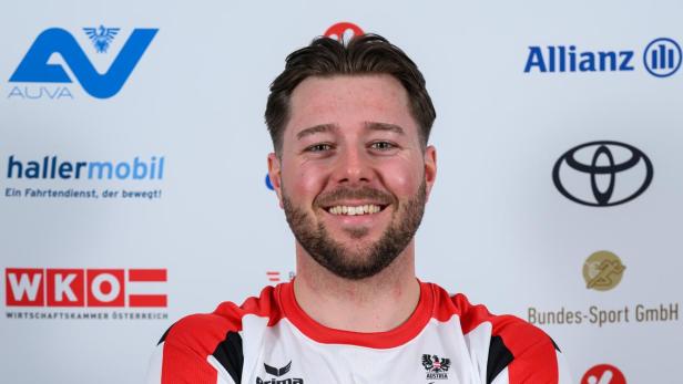 Ein Mann mit verschränkten Armen trägt ein rot-weißes Sporttrikot und lächelt vor einer Sponsorenwand.