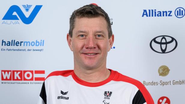 Oliver Dreier: Ein Mann im rot-weißen Sporttrikot mit österreichischem Wappen steht vor einer Sponsorenwand und lächelt.