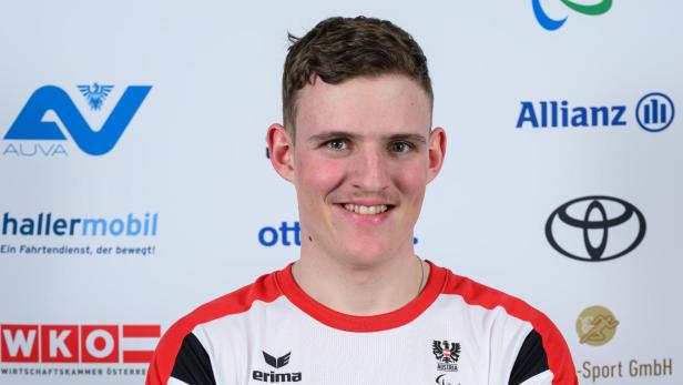 Johannes Aigner: Junger Mann im weißen Sporttrikot mit roten Akzenten steht lächelnd mit verschränkten Armen vor Sponsorenlogos.