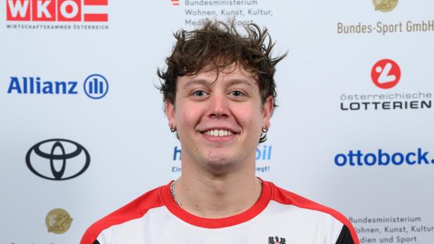 Sebastian Rigby: Junger Mann im Rollstuhl mit kurzem, lockigem Haar trägt ein weißes Sporttrikot mit roten Akzenten und lächelt vor Sponsorenlogos.